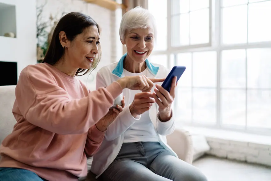Twee mensen zitten samen op een bank en kijken naar een blauwe smartphone voor een groot raam.