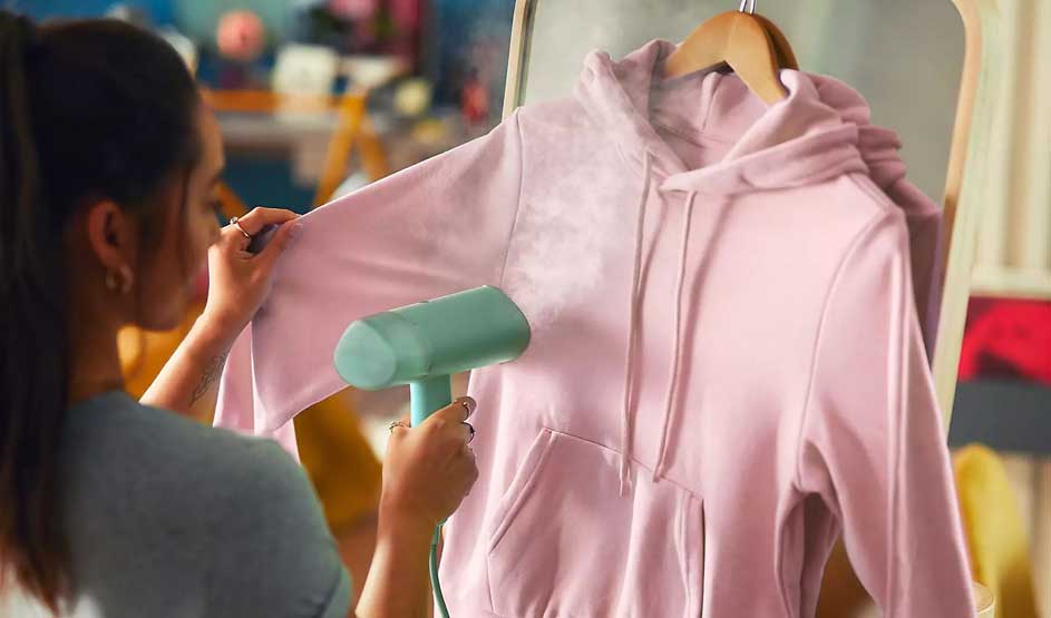 Une personne défroisse un sweat à capuche rose sur cintre, tandis que le tissu devient plus lisse dans une ambiance chaleureuse.