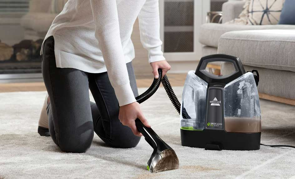 Nettoyage de tapis beige avec un appareil portable noir et transparent. Un canapé gris est visible en arrière-plan.