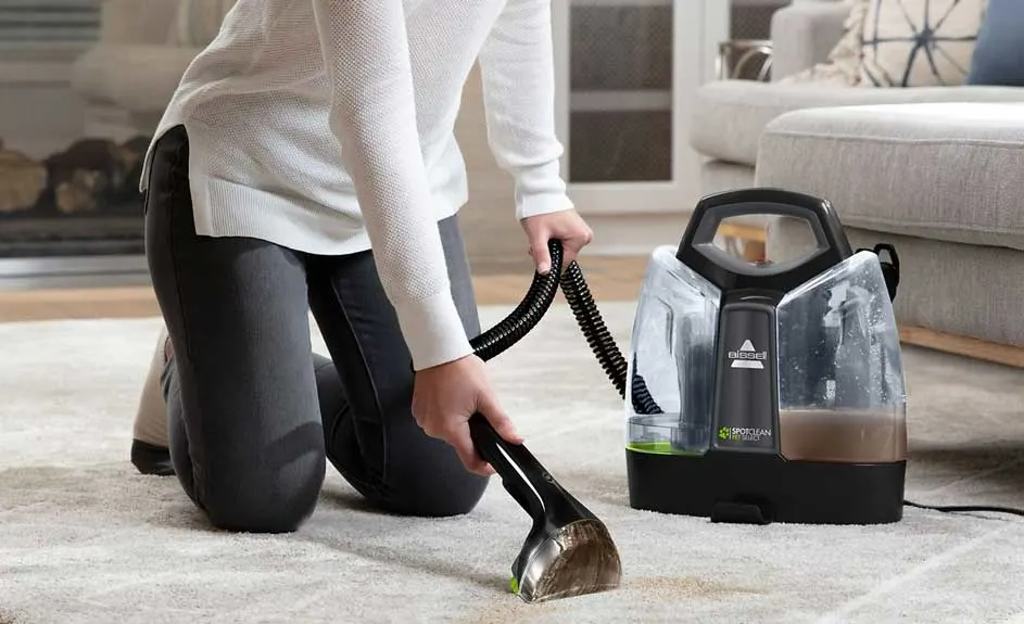 Nettoyage de tapis beige avec un appareil portable noir et transparent. Un canapé gris est visible en arrière-plan.