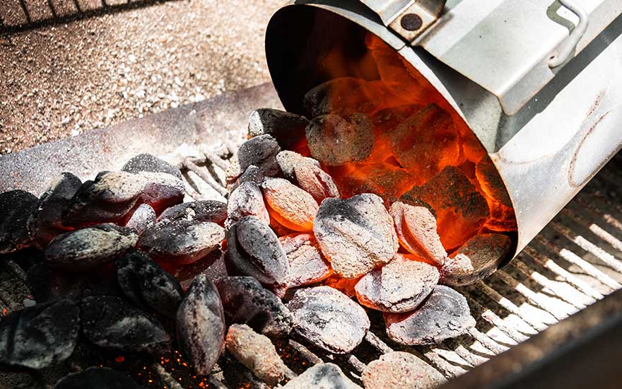 Un contenant en métal verse des briquettes de charbon incandescentes sur une grille de barbecue.