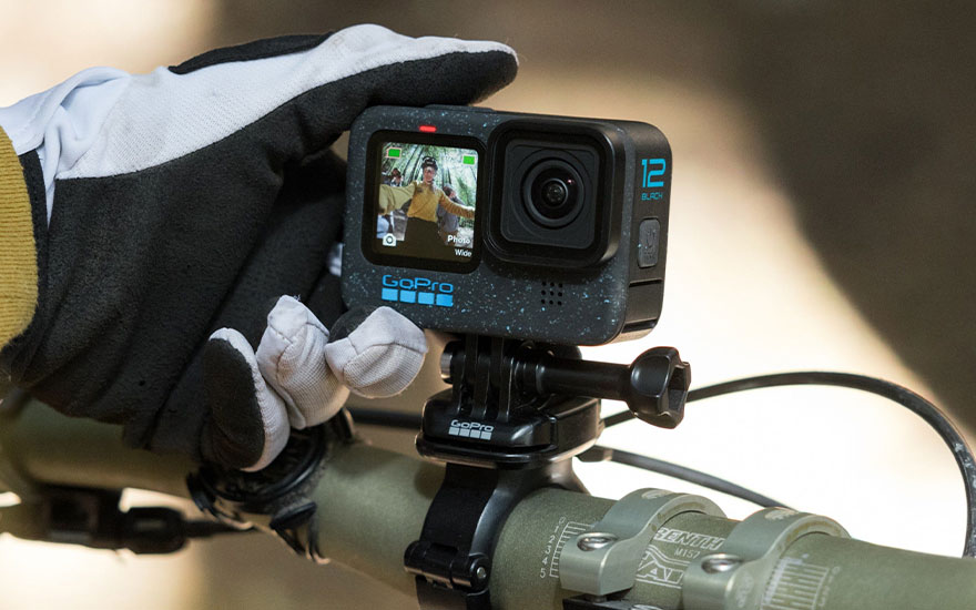 Black GoPro camera mounted on a green bicycle handlebar with a gloved hand nearby.