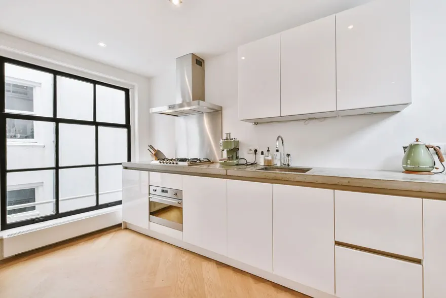 Witte keuken met houten vloer, groot raam en roestvrijstalen afzuigkap.