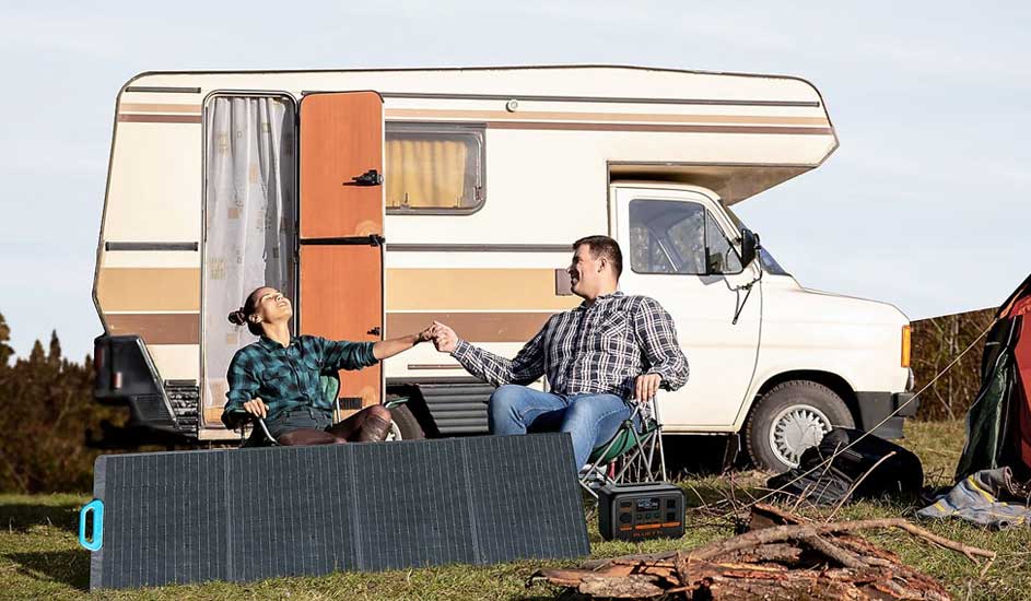 Deux personnes sont assises devant un camping-car avec une station d'énergie BLUETTI et un panneau solaire, main dans la main près d’un feu.