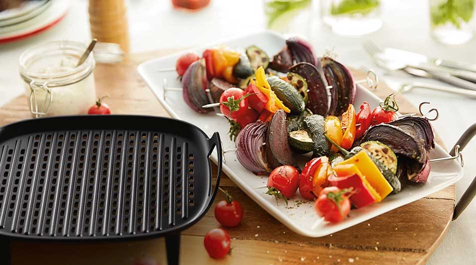 Une poêle à griller noire est à côté d'une assiette blanche de brochettes de légumes colorées sur une planche à découper en bois.