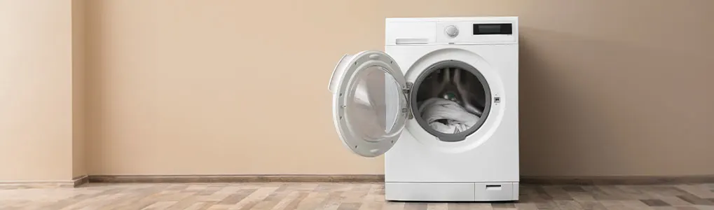 Witte wasmachine met open deur tegen een beige muur, op een houten vloer. Er zit wasgoed in de trommel.