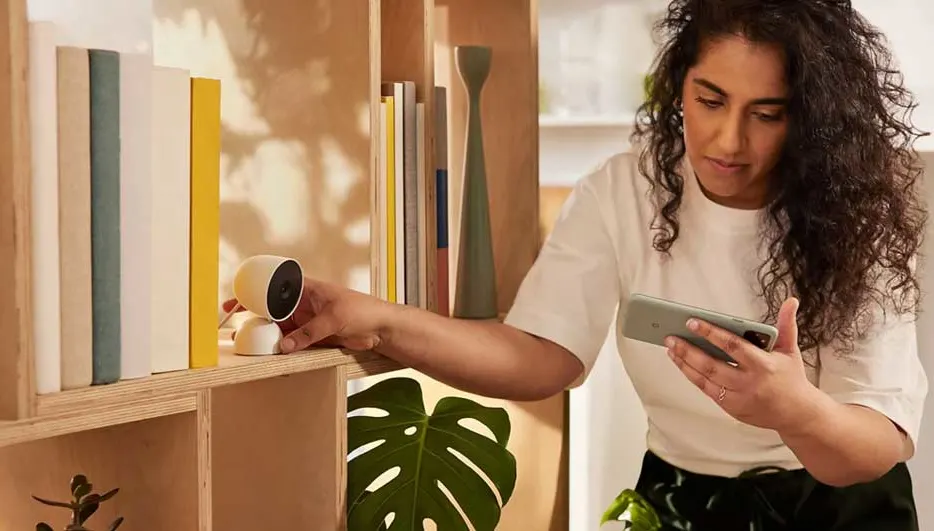 Une étagère en bois contient des livres colorés, une caméra blanche et une plante verte. Un smartphone est tenu à droite.