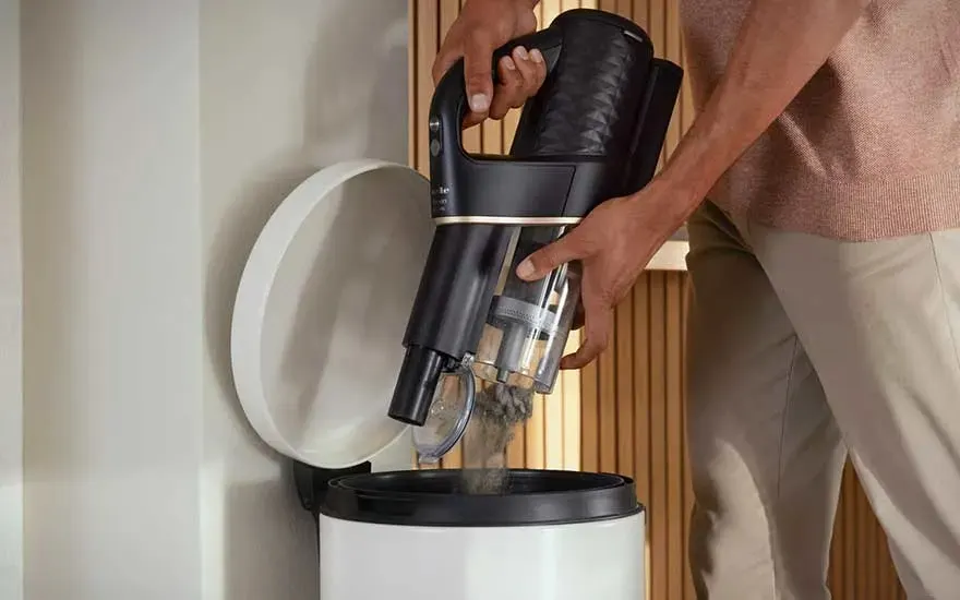 Une personne vide un aspirateur noir dans une poubelle blanche avec un couvercle ouvert.