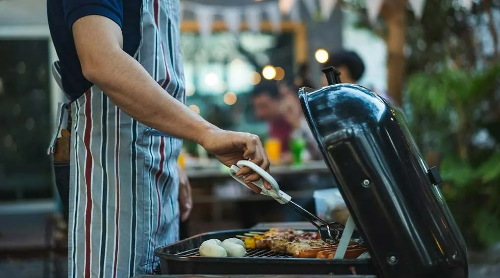 A person in a striped apron uses tongs to grill food on a black grill outdoors.