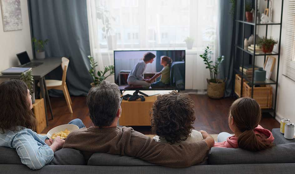Een gezin kijkt samen televisie in een lichte woonkamer met planten, rekken en grote ramen.