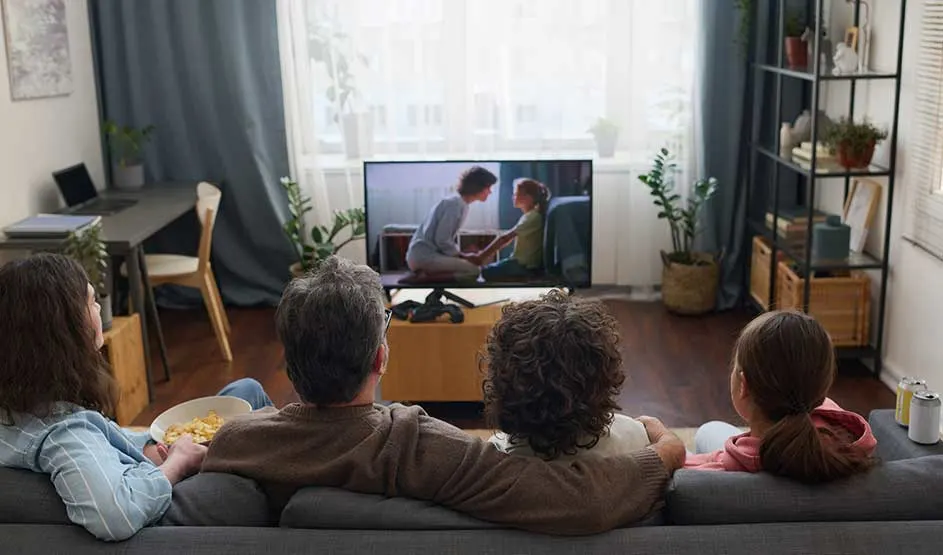 Une famille regarde la télévision ensemble dans un salon lumineux avec plantes, étagères et grandes fenêtres.