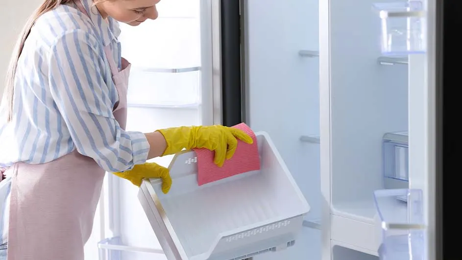 Une personne nettoie un bac de frigo blanc avec un chiffon rose. Elle porte des gants jaunes. L'intérieur du frigo est vide.