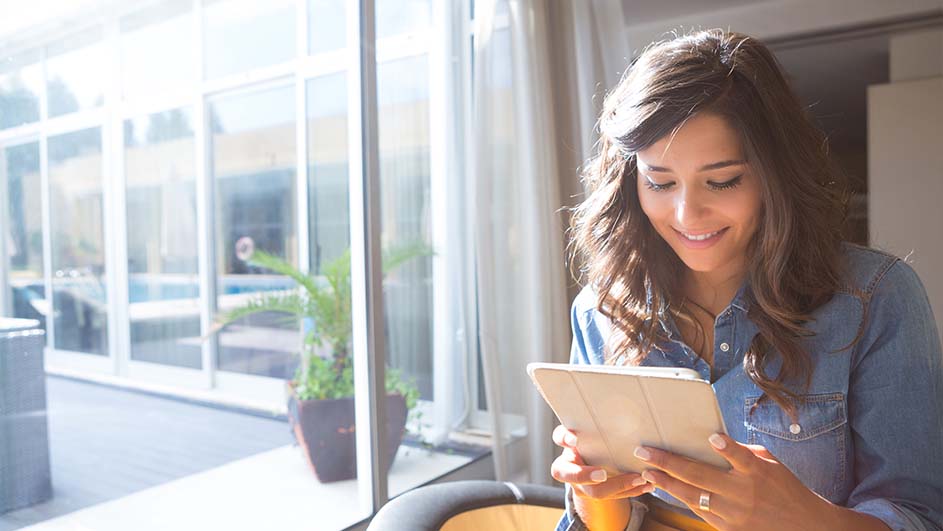 Une tablette est tenue devant une fenêtre lumineuse. Dehors, on aperçoit des plantes en pot et une véranda blanche.