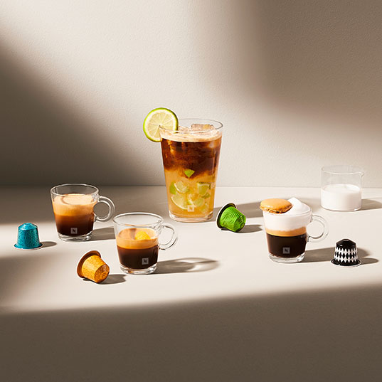 Diverse koffiedranken en capsules op een licht oppervlak. Espressokopjes, een ijskoffie met limoen, en melk in een glas.