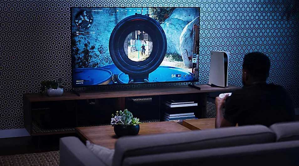 Un écran TV affiche un jeu vidéo avec un viseur. Une console blanche est à côté. Un canapé gris est au premier plan.