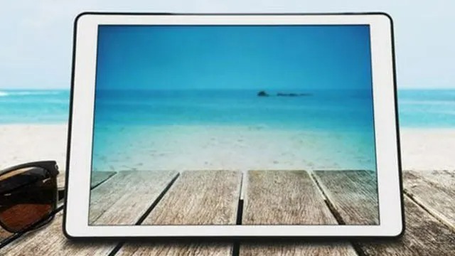 Een witte tablet ligt op een houten tafel met een foto van een blauwe zee en strand erop. Een zonnebril ligt ernaast.