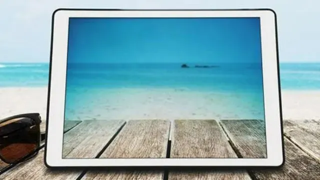 Een witte tablet ligt op een houten tafel met een foto van een blauwe zee en strand erop. Een zonnebril ligt ernaast.