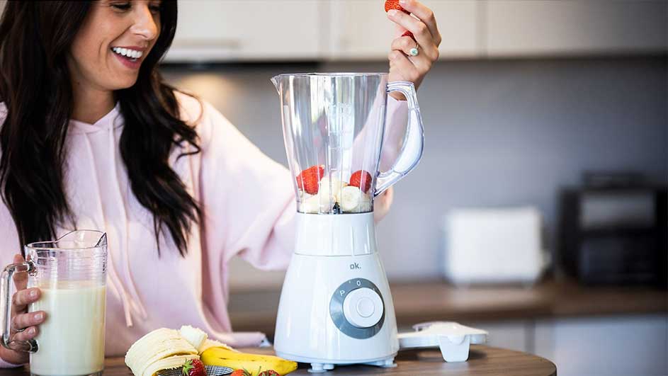 Une fraise est ajoutée à un mixeur contenant des bananes. Un pichet de liquide blanc est à côté.