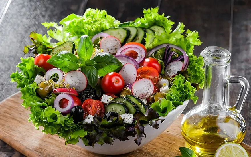 Un bol blanc rempli de salade mixte avec des tomates, des concombres, des oignons rouges et de la laitue. Une bouteille d'huile sur le côté.