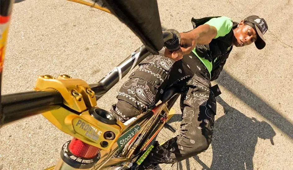 Zicht op een gouden fietsstuur met zwarte handvatten, een zwart frame en een bandana-patroon op de broek van de fietser.