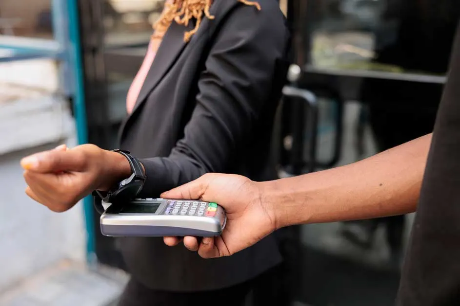 Een dame betaalt contactloos met een horloge. Een hand houdt een betaalterminal vast. Achtergrond: een winkelruit en een deur.