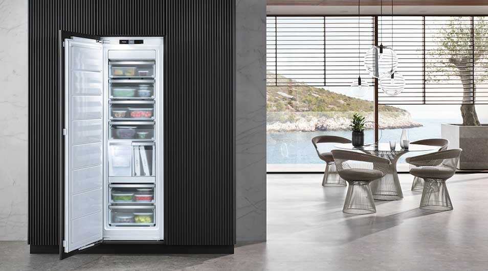 Een openstaande witte koelkast gevuld met voedsel, naast een eettafel met stoelen en een uitzicht op zee.