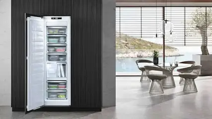 Frigo blanc ouvert intégré dans un mur noir, révélant des tiroirs remplis. Table à manger et vue sur la mer en arrière-plan.