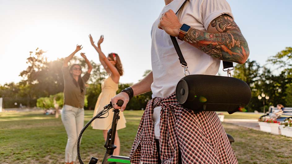 Een persoon met een fiets en speaker staat in een park. Twee andere personen staan op de achtergrond met hun armen omhoog.
