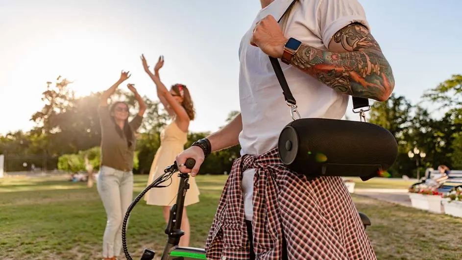 Een persoon met een fiets en speaker staat in een park. Twee andere personen staan op de achtergrond met hun armen omhoog.