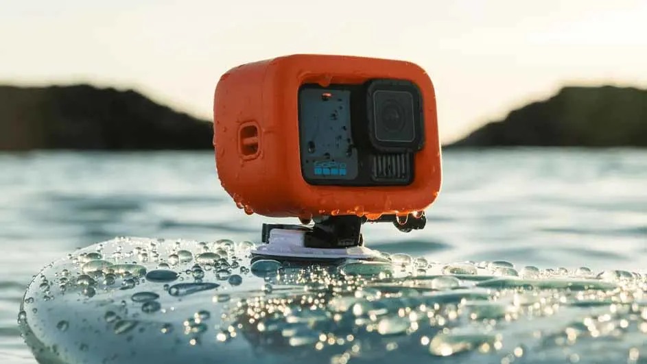 An orange action camera on a mount sits on a wet surface, with water and hills in the background.