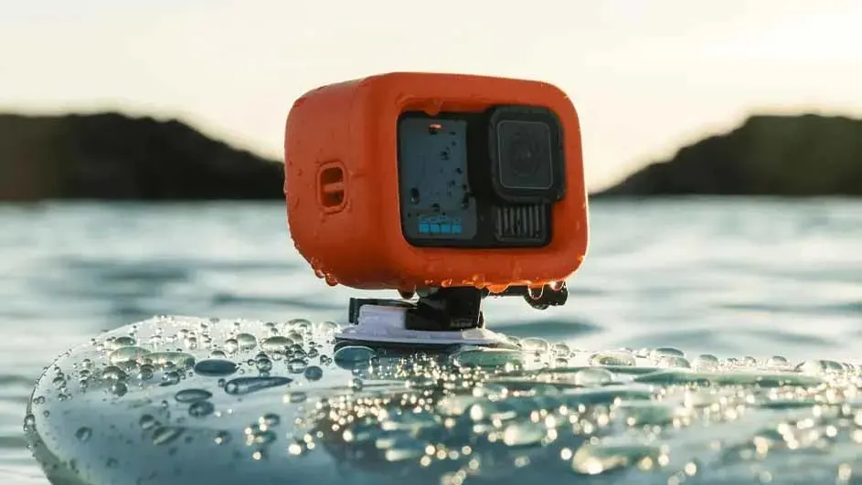 An orange action camera on a mount sits on a wet surface, with water and hills in the background.
