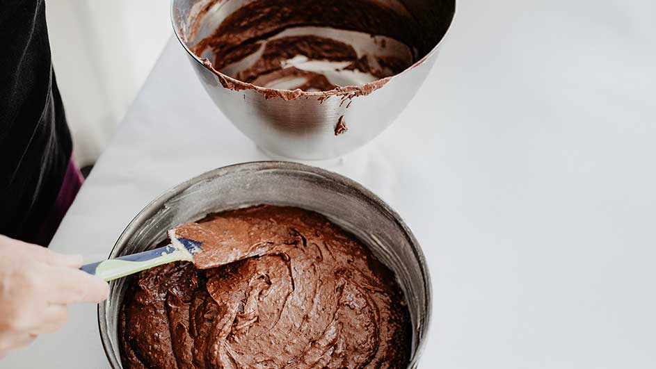 Un moule rond en métal contient de la pâte à gâteau au chocolat. Une spatule étale la pâte. Un autre bol avec de la pâte est visible.