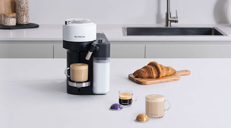 Black and white coffee maker with milk frother, croissant on wood board, coffee cups, capsules on white counter.