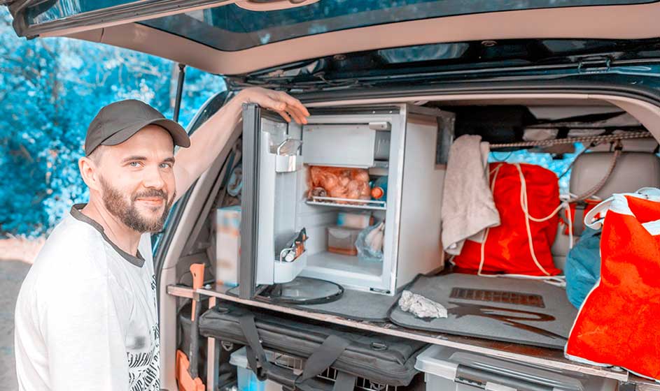 Een glimlachende man opent de achterklep van een auto met een compacte koelkast. Er zijn groenten, drankjes, tassen en kampeerspullen zichtbaar.