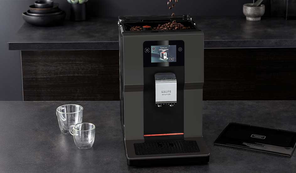 A black automatic coffee machine stands on a dark worktop with coffee beans on top. Two empty glasses and a black booklet are placed beside it.