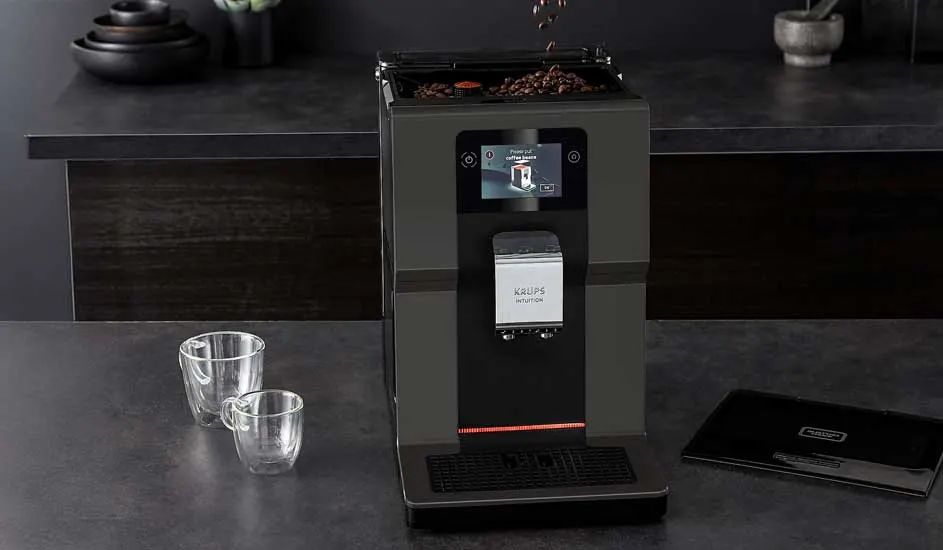 A black automatic coffee machine stands on a dark worktop with coffee beans on top. Two empty glasses and a black booklet are placed beside it.