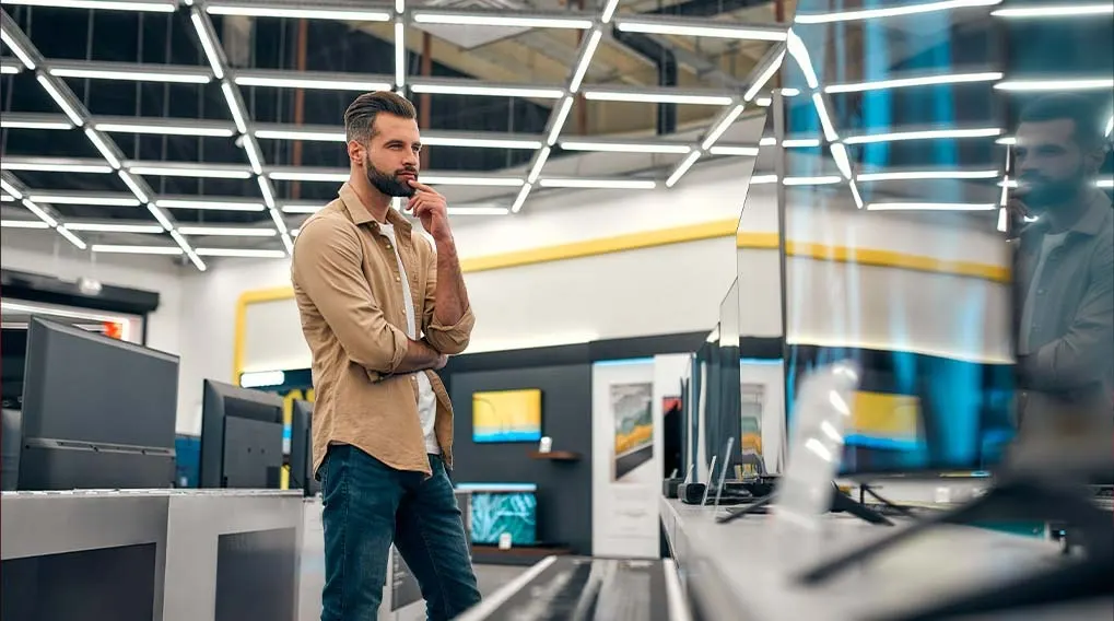 Dans un magasin, des télévisions sont exposées sur des étagères sous un plafond lumineux à motifs géométriques.