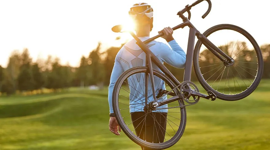 Un cycliste porte un vélo noir sur l'épaule dans un champ verdoyant avec des arbres au loin sous un soleil éclatant.