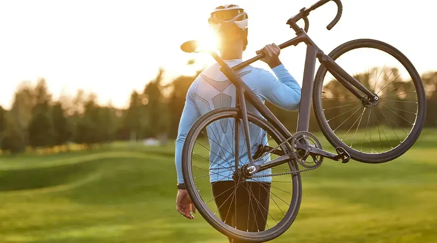Un cycliste porte un vélo noir sur l'épaule dans un champ verdoyant avec des arbres au loin sous un soleil éclatant.