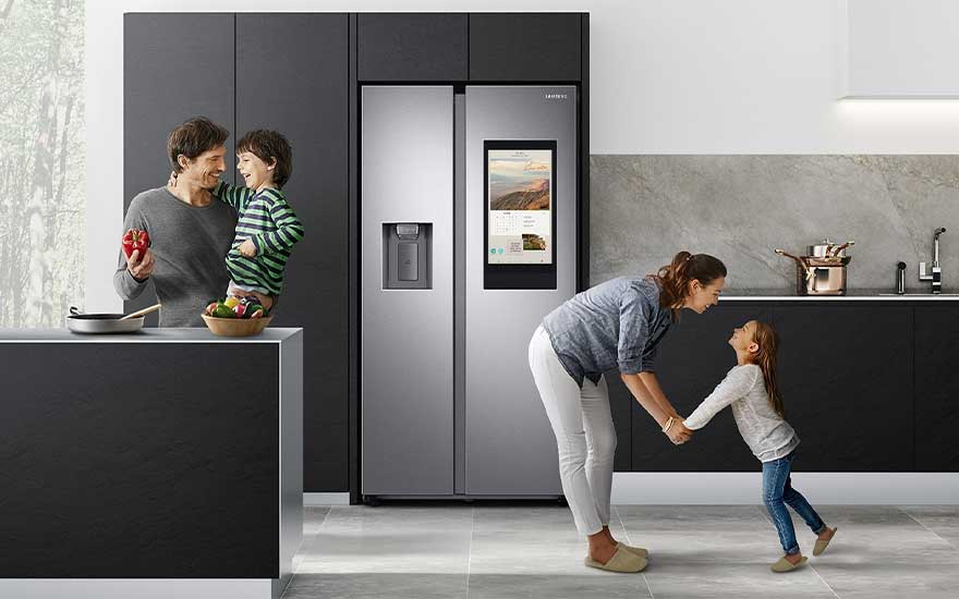 Cuisine moderne avec frigo argenté à écran, meubles noirs, plan de travail blanc. Ustensiles et légumes présents.