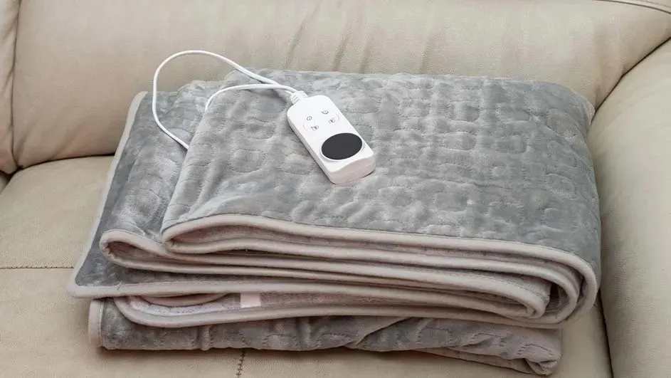 Een grijze, opgevouwen elektrische deken ligt op een beige sofa met een witte afstandsbediening.