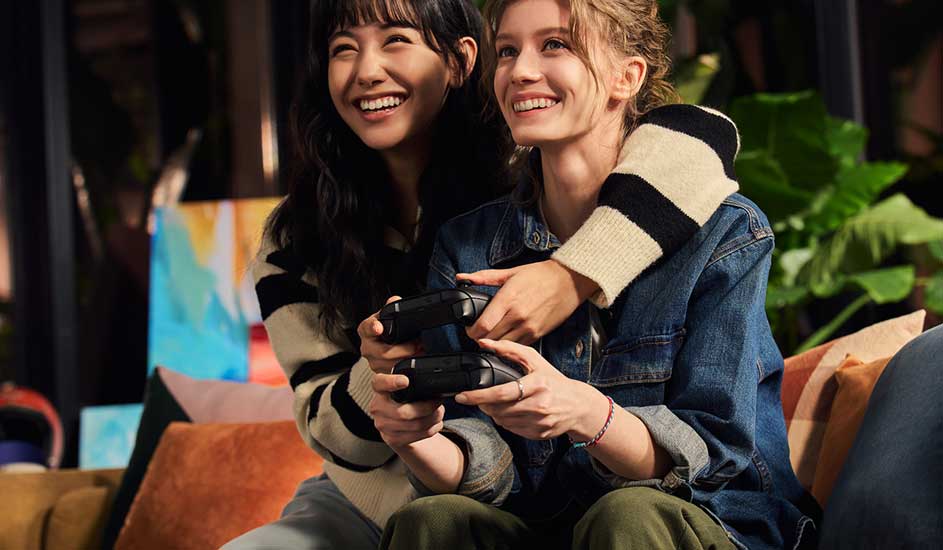 Deux personnes sur un canapé jouent à un jeu vidéo avec une manette noire, dans un intérieur avec des plantes.