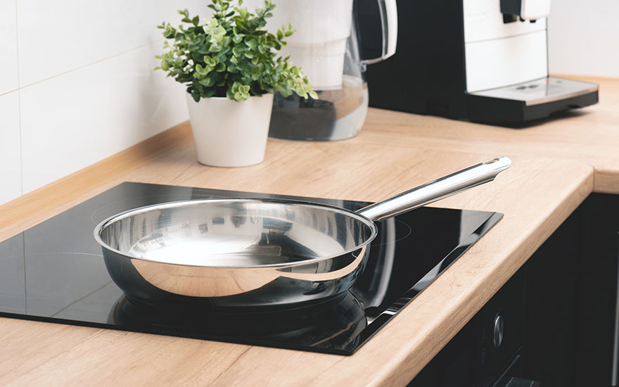 Een zilveren pan staat op een zwarte kookplaat op een houten aanrecht, met een plant en koffiezetapparaat op de achtergrond.