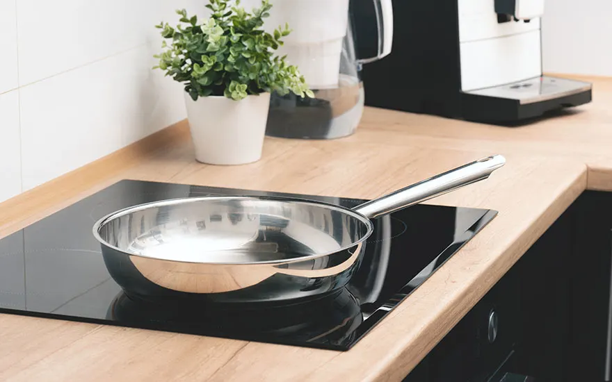 Een zilveren pan staat op een zwarte kookplaat op een houten aanrecht, met een plant en koffiezetapparaat op de achtergrond.