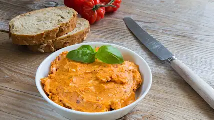 Bol blanc avec tartinade orange, basilic, pain, tomates et couteau sur table en bois.