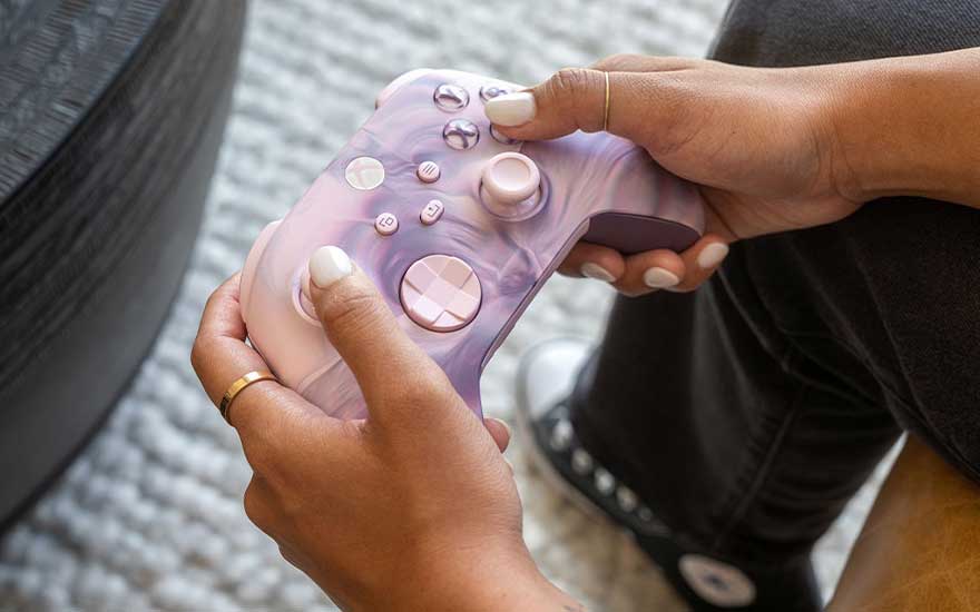 Une manette de jeu rose et violet est tenue par deux mains, avec un canapé sombre en arrière-plan.