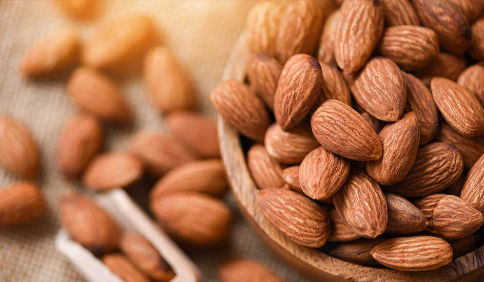 Un bol en bois rempli d'amandes brunes, avec d'autres amandes éparpillées sur une surface texturée. Une cuillère en bois est visible.
