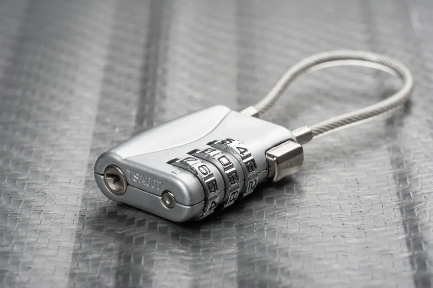 Een zilverkleurig TSA-slot met een flexibele kabel ligt op een zilverkleurige ondergrond. Het slot heeft drie cijferrollen.