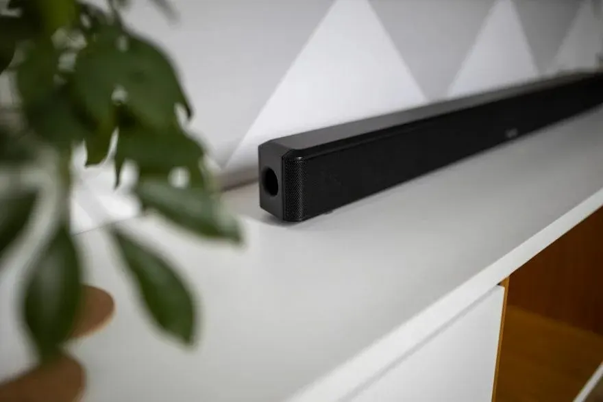 Une barre de son noire repose sur une étagère blanche, avec un motif géométrique gris au mur et des feuilles vertes au premier plan.