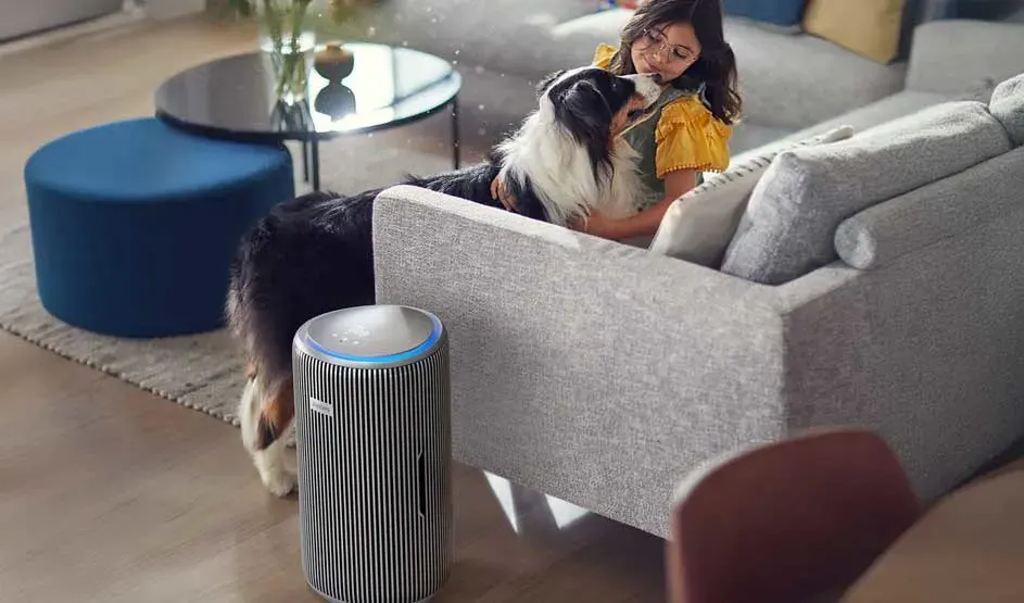 Een meisje op een sofa knuffelt een grote hond, terwijl voor de bank een hoge cilindervormige luchtreiniger staat in een moderne woonkamer.